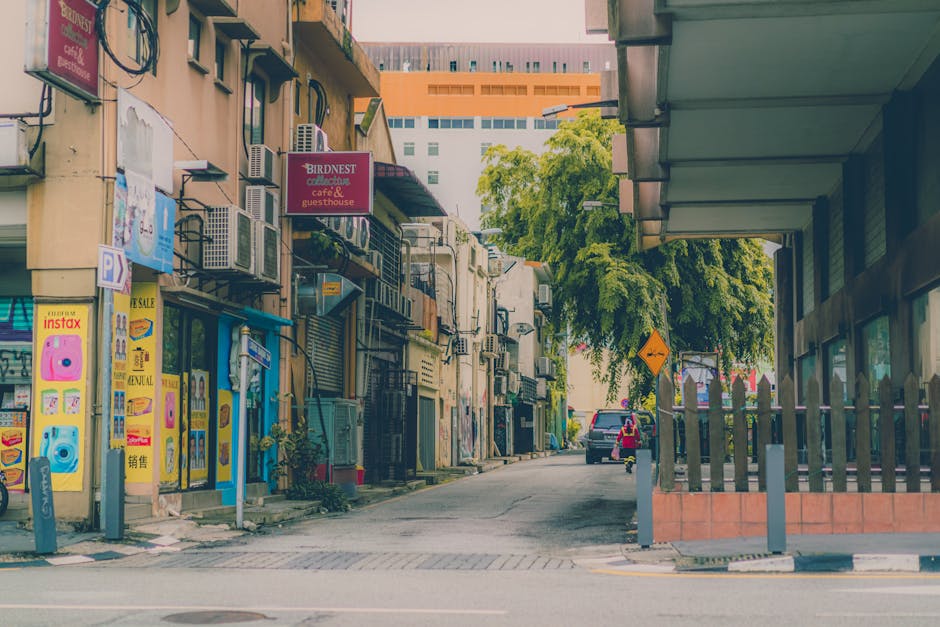 A narrow street in Westcombe Park lined with multi-story buildings featuring exterior walls with air conditioning units and various signage, including a sign for Bird nest cafe and guesthouse, and bright yellow posters advertising an instant camera sale. The street has a small sidewalk with bollards and a parking area with a dark-colored vehicle parked near the curb. A large green tree with full foliage extends partly over the street, providing shade. The scene is well-lit with daylight, showcasing the urban environment used in the context of home relocation or furniture transport involving building access and street logistics. Man with Van Westcombe Park is involved in logistical tasks such as loading or moving furniture and packing materials during a house removal or moving process on a busy street with typical city architecture.