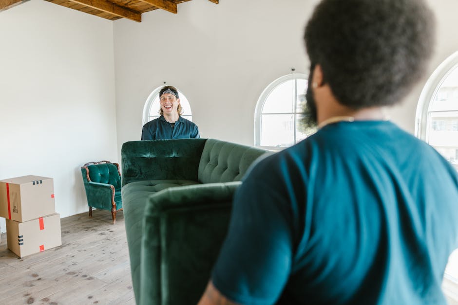 Inside a well-lit room with wooden ceiling beams and white walls, a professional home relocation team from Man with Van Westcombe Park is engaged in a furniture removal process. Two movers, dressed in blue uniforms, are handling a large, green upholstered sofa, which is being carried through the room. One mover, with his back to the camera, is visibly supporting the sofa from the front, while the other, standing near the window, is guiding its movement. In the background, cardboard boxes sealed with red packing tape are stacked on the wooden floor, indicating packing and loading activities. The room features two arched windows allowing natural light to illuminate the space and a small vintage armchair with wooden legs positioned against the wall. The movers are in the process of loading the sofa, likely preparing for transport during a house removal or home relocation service offered by [COMPANY_NAME], supporting smooth furniture transport and packing logistics as part of their removals service in Westcombe Park.