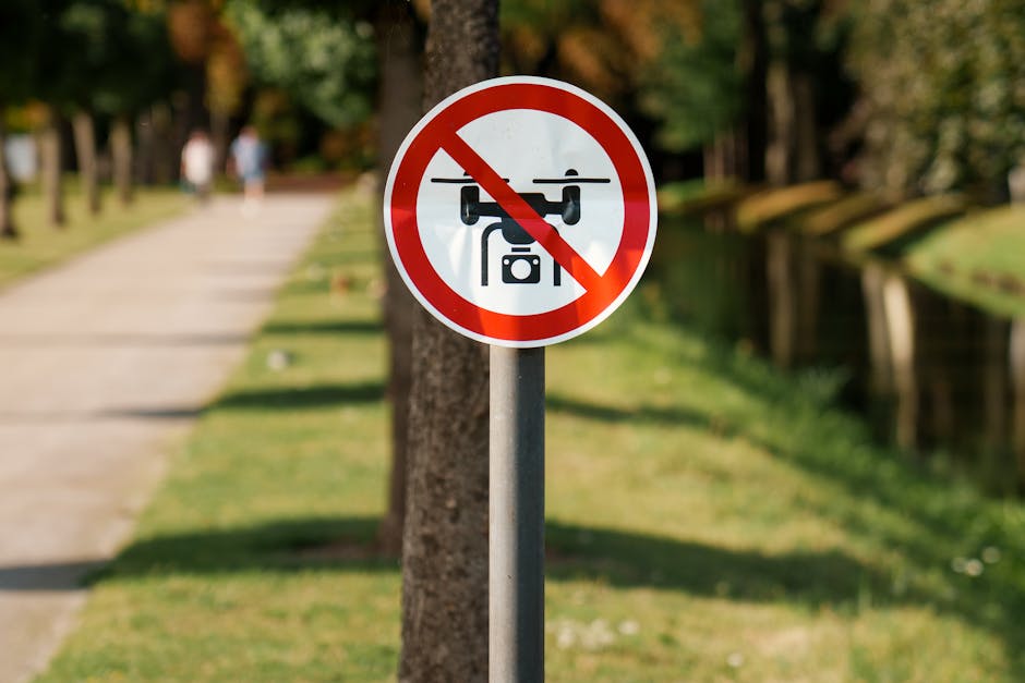 A circular sign with a red border and white background displaying a black drone icon with a red slash through it, indicating no drone flying allowed. The sign is mounted on a metal pole located along a pathway in a park-like setting with a grassy verge, trees, and a wooden barrier in the background. There are a few blurred pedestrians walking along the pathway, and the area is illuminated by natural daylight. This image shows signage relevant to urban regulations, similar to those encountered during house removals or moving activities near public spaces, emphasizing restrictions on drone usage in controlled or residential areas. The scene is captured during daytime with clear weather, highlighting the importance of adhering to local regulations during home relocation processes managed by companies like Man with Van Westcombe Park.