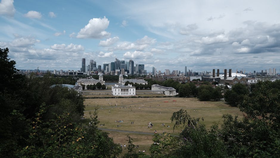 A wide view of Greenwich Park in London under a partly cloudy sky, with lush green trees framing the foreground. In the middle ground, the historic Queen's House with its white facade and classical architecture is visible, surrounded by open grassy areas with visitors sitting and relaxing. In the background, the modern city skyline with tall skyscrapers and office buildings rises prominently, contrasting with the historic park and indicating a city-centre location. The scene illustrates a peaceful urban green space during daytime, with clear visibility of the various architectural styles and natural elements, ideal for activities such as house removals or furniture transport planning with [COMPANY_NAME], who often assist with packing, loading, and relocating household items within this vibrant area.