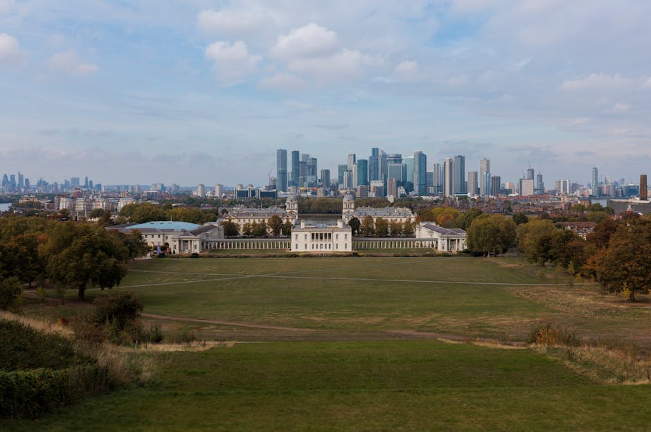 A wide panoramic view of Greenwich Park in London during daytime, with lush green grass, several mature trees on either side, and classical pavilion buildings centrally positioned in the foreground. Beyond the park, the modern London skyline with high-rise office buildings and skyscrapers stretches across the horizon under a partly cloudy sky. The scene captures the contrast between historic park architecture and contemporary urban development, illustrating a typical setting for house removals and furniture transport services such as those provided by Man with Van Westcombe Park, with the park serving as a peaceful green space near busy city areas.