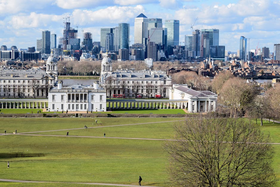 A wide view of Greenwich Park in London under a partly cloudy sky, with lush green trees framing the foreground. In the middle ground, the historic Queen's House with its white facade and classical architecture is visible, surrounded by open grassy areas with visitors sitting and relaxing. In the background, the modern city skyline with tall skyscrapers and office buildings rises prominently, contrasting with the historic park and indicating a city-centre location. The scene illustrates a peaceful urban green space during daytime, with clear visibility of the various architectural styles and natural elements, ideal for activities such as house removals or furniture transport planning with [COMPANY_NAME], who often assist with packing, loading, and relocating household items within this vibrant area.