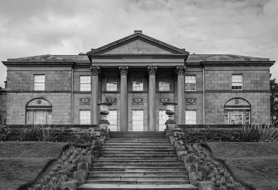 A close-up view of the exterior facade of a historic building featuring large, classical columns with fluted designs supporting a triangular pediment, set against a clear blue sky with a few wispy clo