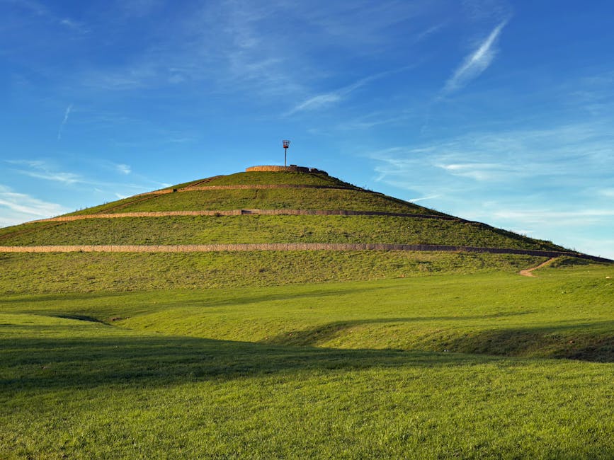A large grassy hill with neatly maintained terraced steps leading to an observation tower at the summit, under a bright blue sky with wispy white clouds. The foreground consists of expansive green lawns with gentle undulations, creating a scenic landscape. The hill's terraced paths are visible, running from the base to the top, providing access for visitors. At the top, a small structure or platform supports the observation tower, which is equipped with a red and white weather station or antenna. The natural environment appears well-kept, with no visible buildings or urban elements, suggesting a park or open recreational area. This landscape is suitable for outdoor activities such as walking or enjoying panoramic views, and it exemplifies a typical scenic feature that could be encountered during a home relocation or moving process in a landscaped or rural setting. Occasionally, a removal company such as Man with Van Westcombe Park might assist in transporting belongings through or around such areas, ensuring smooth logistics during a house move.