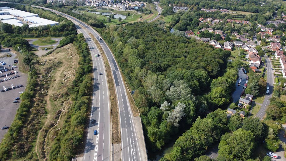Aerial view of a highway running alongside a densely wooded area with tall green trees and a small river visible through the foliage. To the right of the wooded area, there is a residential neighbourhood with rows of houses, streets, and parked cars. On the left side, a commercial parking lot with numerous cars is situated near a large building. The scene is captured during daylight, with clear skies overhead. This image highlights the proximity of natural landscape to urban development, which may be relevant to house removals and relocation logistics provided by Man with Van Westcombe Park.