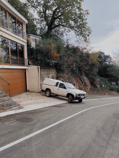 A white van parked on a concrete driveway outside a residential property, with the front of the van facing the street. The driveway is adjacent to a modern house featuring large glass windows and sliding doors with metal railings on a balcony. The van is positioned near a set of stone steps leading to the house entrance, and a wooden garage door is visible on the left side of the image. Behind the van, there is a retaining wall and a slope covered with trees and bushes, indicating a hillside environment. The scene is captured during daylight with overcast skies, suggesting suitable conditions for a home relocation or furniture transport process as part of a professional removals service by Man with Van Westcombe Park, who manages loading and moving activities efficiently in residential settings.