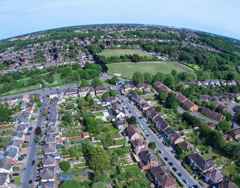 Aerial view of a highway running alongside a densely wooded area with tall green trees and a small river visible through the foliage. To the right of the wooded area, there is a residential neighbourhood with rows of houses, streets, and parked cars. On the left side, a commercial parking lot with numerous cars is situated near a large building. The scene is captured during daylight, with clear skies overhead. This image highlights the proximity of natural landscape to urban development, which may be relevant to house removals and relocation logistics provided by Man with Van Westcombe Park.