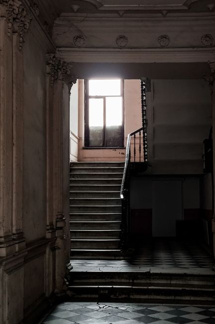 Inside an old, dimly lit staircase area of a Georgian-style home in Westcombe Park, with marble steps leading upward toward a window that allows natural light to illuminate the space. The staircase has a black metal handrail on the right side, and the surrounding walls feature ornate decorative mouldings and wood paneling. The floor at the base of the stairs is tiled with black and white checkered patterns, and the overall scene captures a historic, somewhat abandoned interior environment. The image's context relates to home relocation or moving services, with potential considerations for safely navigating stairwells during furniture transport or packing and moving procedures, as offered by Man with Van Westcombe Park.