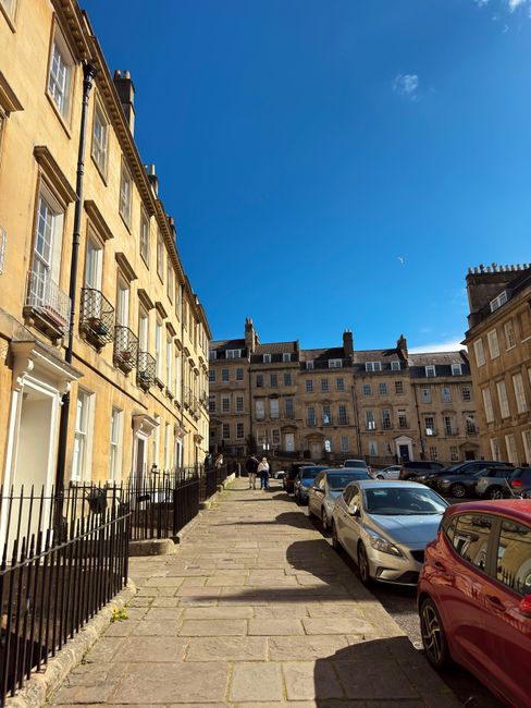 Image of a residential street in Westcombe Park with a row of classic Georgian-style terraced houses on the left, featuring cream-colored facades, black metal balcony railings, and sash windows. The pavement, made of large stone slabs, runs along the front of the homes, with a black metal fence separating the sidewalk from the front gardens. On the right side, several cars of various makes and models are parked along the curb, including a red hatchback, a silver sedan, and other vehicles. The sky above is clear and blue, with bright sunlight casting shadows on the pavement and buildings. In the background, a few people are seen walking towards a small group near the end of the street, suggesting a typical city neighbourhood. This scene could represent an area where professional furniture transport and home relocation services, such as those offered by Man with Van Westcombe Park, might be required for moving purposes within Georgian Westcombe Park homes, involving careful planning of loading and unloading processes in a residential setting.