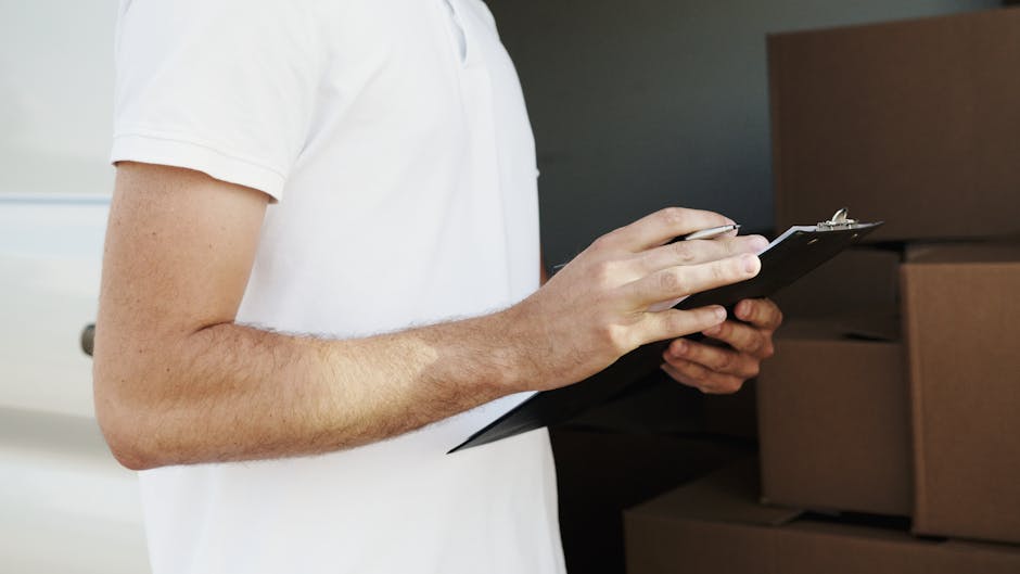 Close-up of a man wearing a white T-shirt, holding and writing on a clipboard with a pen, standing inside a property near stacked cardboard boxes wrapped in clear plastic. The boxes are positioned on the floor against a dark-colored wall, indicating the packing and moving process as part of home relocation efforts. The lighting is natural, highlighting the man's arms and the packing materials, with a focus on the preparation stage of furniture transport and packing for a same-day removal service. The scene reflects an organized approach to loading and managing belongings during a professional move, supported by Man with Van Westcombe Park's removals service.