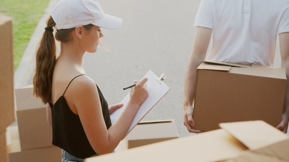 Close-up of a man wearing a white T-shirt, holding and writing on a clipboard with a pen, standing inside a property near stacked cardboard boxes wrapped in clear plastic. The boxes are positioned on the floor against a dark-colored wall, indicating the packing and moving process as part of home relocation efforts. The lighting is natural, highlighting the man's arms and the packing materials, with a focus on the preparation stage of furniture transport and packing for a same-day removal service. The scene reflects an organized approach to loading and managing belongings during a professional move, supported by Man with Van Westcombe Park's removals service.