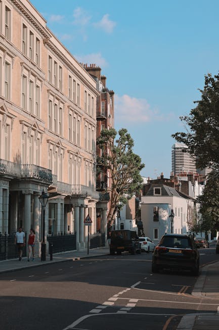 A quiet residential street in Westcombe Park during daytime, featuring a row of elegant, cream-coloured Georgian-style houses with large windows, decorative cornices, and black iron balconies. The pavement is lined with tall, leafy trees casting shadows on the roadway, and several parked cars are visible along the curb, including a black van near the entrance of a property. In the background, modern high-rise buildings contrast with the historic architecture. The scene includes a few pedestrians walking on the sidewalk, possibly residents or visitors. The sky is clear with scattered clouds, and the overall setting suggests a peaceful neighbourhood suitable for home relocation or local house removals, with [COMPANY_NAME]'s services supporting furniture transport and packing processes during a move in the Westcombe Park area.