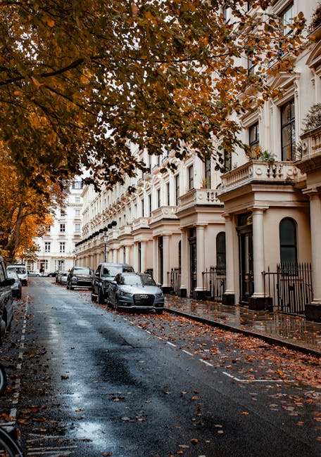 A residential street in Westcombe Park lined with white-painted Victorian-style terraced houses featuring bay windows, decorative moldings, and wrought iron railings at the front. The cobblestone street is wet, with puddles reflecting the overcast sky, and scattered fallen autumn leaves covering the pavement. Parked cars, including a black SUV and a silver sedan, are positioned alongside the curb. A large tree with orange and brown foliage overhangs the street, its branches extending over the parked vehicles and sidewalk. This scene captures the quiet atmosphere of a house removal in progress, with no visible moving equipment or personnel, but the environment suggests preparation for home relocation or furniture transport involving packing and loading nearby. The context aligns with professional removals services such as those offered by Man with Van Westcombe Park, operating within this area.