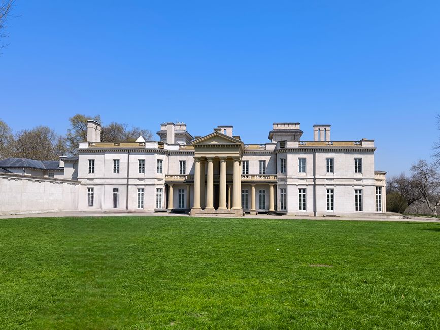 A large, historic white manor house with classical architectural features, including a prominent portico supported by six tall, beige columns, situated behind a wide, well-maintained green lawn. The building has multiple stories with numerous evenly spaced windows, and the roofline is adorned with decorative chimney stacks and parapets. The area in front of the house appears to be a driveway or loading zone, with a clear blue sky overhead and some sparse trees visible in the background. This scene is indicative of a property that could be involved in a home relocation or furniture transport project, with the exterior environment suggesting a setting where professional removals, such as those from Man with Van Westcombe Park, might facilitate moving and packing services.
