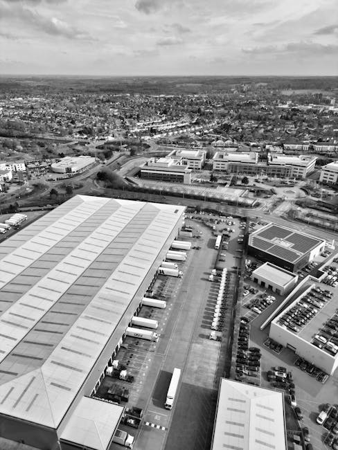 Aerial black-and-white photograph of a large industrial warehouse with a spacious loading bay, where multiple trucks are parked alongside the building. The warehouse's flat roof features evenly spaced skylights, and large doors are open for loading and unloading. Several cardboard boxes, wrapped in plastic or fabric blankets, are arranged near the trucks, indicating preparations for or completion of a home relocation or furniture transport process. The scene includes a paved yard area, with trolleys and other equipment used for moving items, and is surrounded by additional commercial buildings and parking spaces. Beyond the industrial complex, there is an extensive view of a cityscape and open land in the distance under overcast skies, with natural light illuminating the scene. Such environments are typical for commercial moving operations managed by companies like Man with Van Westcombe Park, supporting packing, loading, and transportation for house removals and relocation services.
