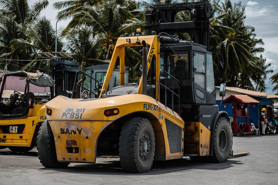 A large yellow SANY forklift with black accents is parked outdoors in a busy area, surrounded by palm trees and structures including a red-roofed building. The forklift's cabin is enclosed with glass windows, and it has a long lifting arm extending forward, with a pair of black forks resting on the ground. The vehicle features thick, heavy-duty tires and visible branding and operational markings. Nearby, other construction or industrial equipment is present, including a partially visible yellow piece of machinery, indicating a setting related to construction, logistics, or equipment transportation, where loading, unloading, or logistics activities are taking place, consistent with house removal or furniture transport processes handled by specialists like Man with Van Westcombe Park.