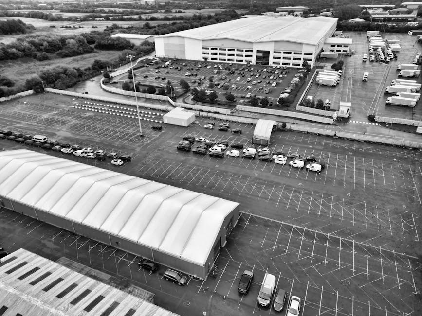 Aerial black-and-white photograph of a large industrial warehouse with a spacious loading bay, where multiple trucks are parked alongside the building. The warehouse's flat roof features evenly spaced skylights, and large doors are open for loading and unloading. Several cardboard boxes, wrapped in plastic or fabric blankets, are arranged near the trucks, indicating preparations for or completion of a home relocation or furniture transport process. The scene includes a paved yard area, with trolleys and other equipment used for moving items, and is surrounded by additional commercial buildings and parking spaces. Beyond the industrial complex, there is an extensive view of a cityscape and open land in the distance under overcast skies, with natural light illuminating the scene. Such environments are typical for commercial moving operations managed by companies like Man with Van Westcombe Park, supporting packing, loading, and transportation for house removals and relocation services.