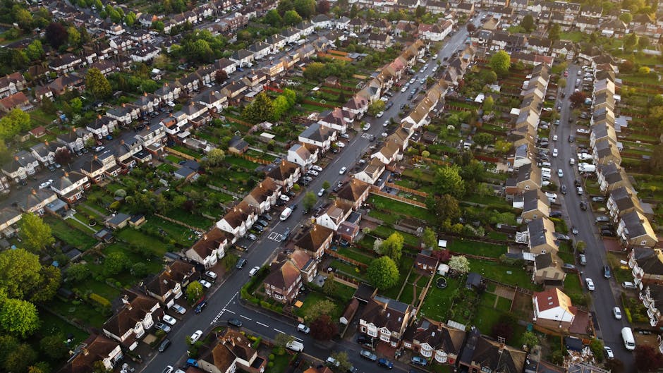 Aerial view of a residential neighbourhood on a sunny day, showing rows of terraced houses with small front gardens, driveways, and parked cars along the streets. The houses are made of brick with pit